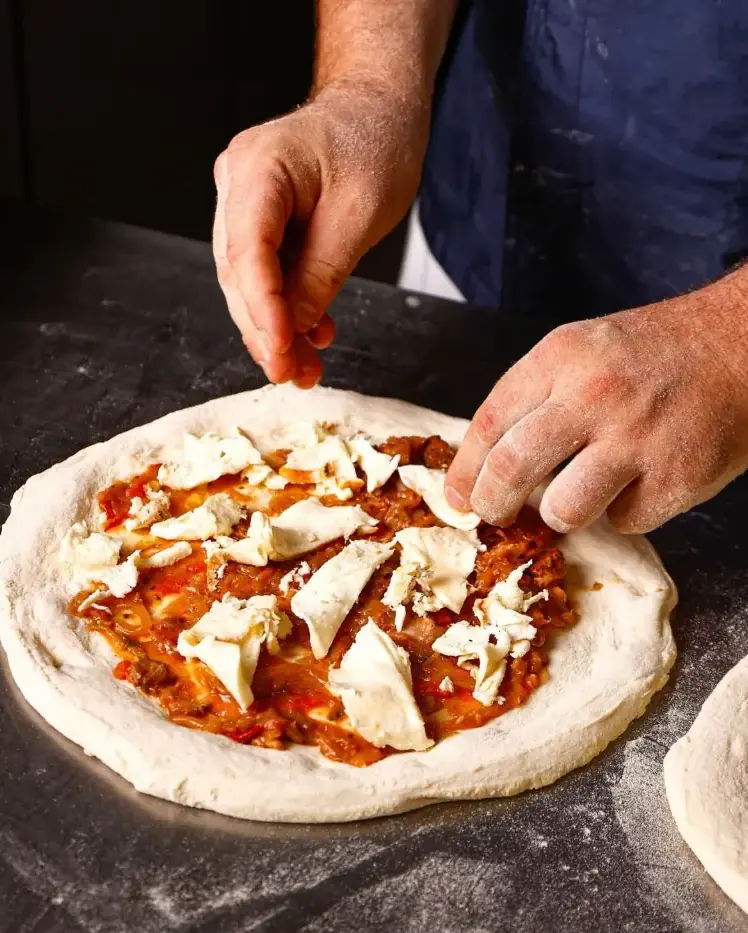 Le mani del pizzaiolo condiscono una pizza appena stesa sul banco